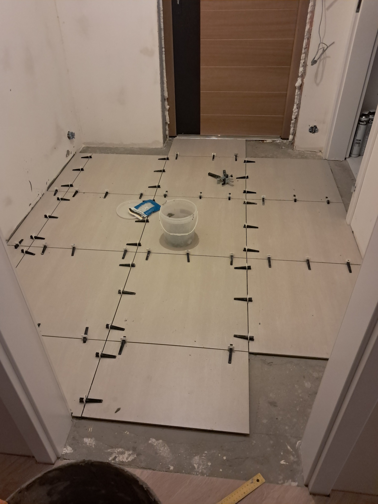 Jasne płytki podłogowe w trakcie układania, z krzyżykami dystansowymi, widok z góry. Częściowo ułożone, z narzędziami i wiadrem w tle. Wnętrze w trakcie prac wykończeniowych.