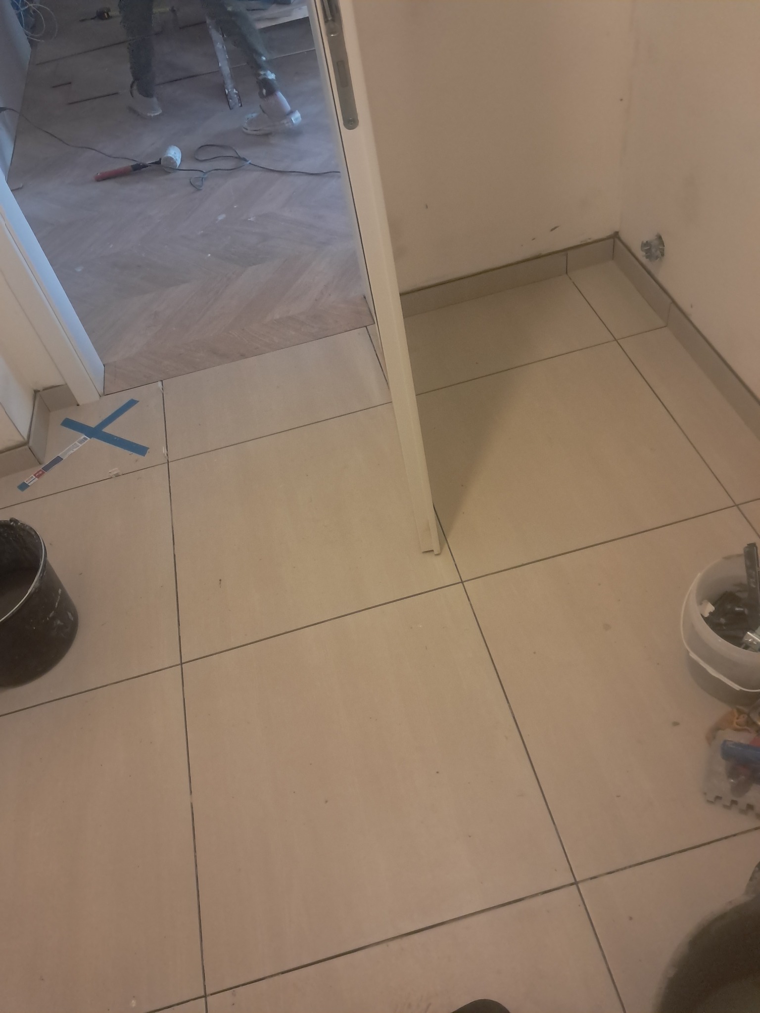 Ułożone beżowe płytki podłogowe w przedpokoju, widoczne narzędzia i fragment podłogi w innym wzorze. Praca w toku, detale wykończeniowe.