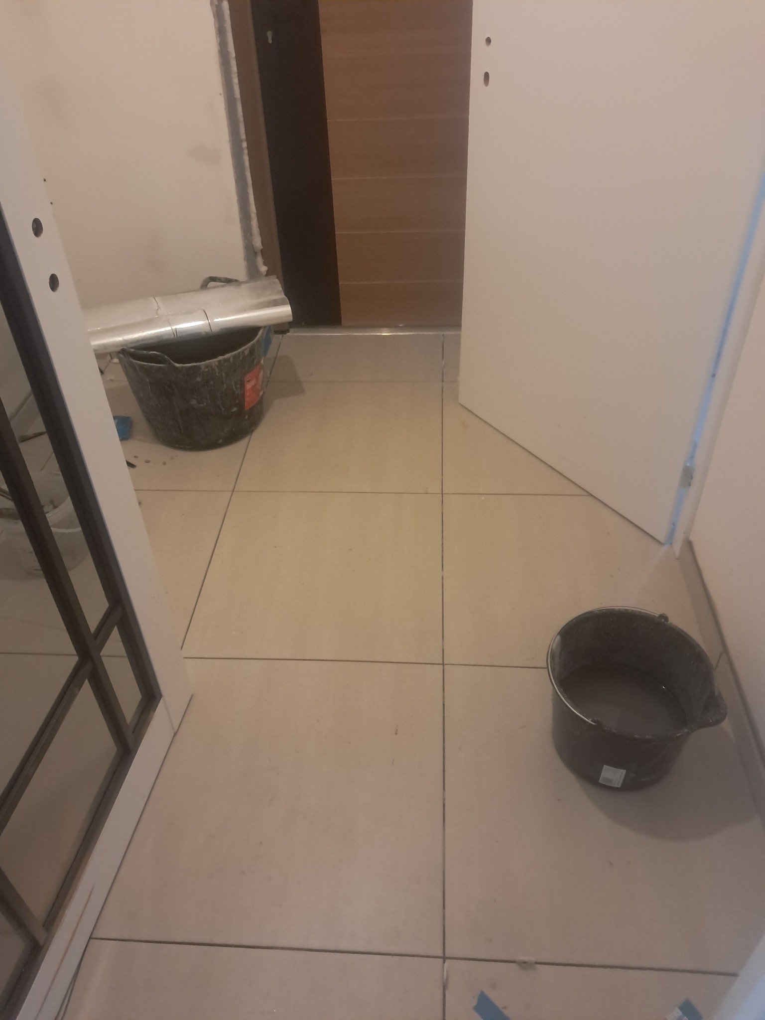 Ułożone, jasne płytki podłogowe w trakcie prac wykończeniowych. Widoczne dwa wiadra z wodą i fragmenty drzwi. Ujęcie z góry, blisko podłogi.