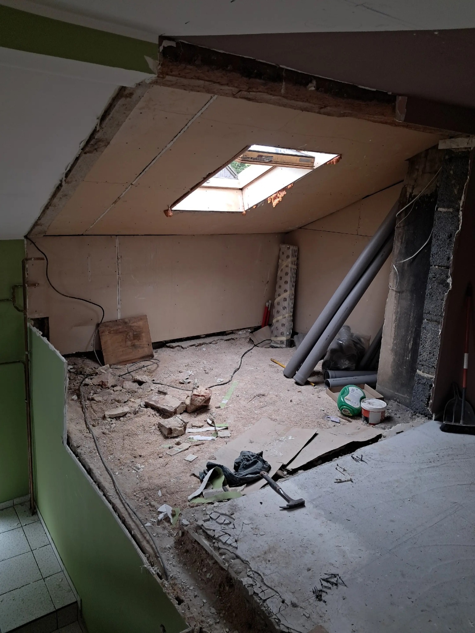 Wnętrze poddasza w trakcie demontażu: odsłonięte płyty kartonowo-gipsowe, gruz na podłodze, widoczne rury i narzędzia. Okno dachowe w suficie.