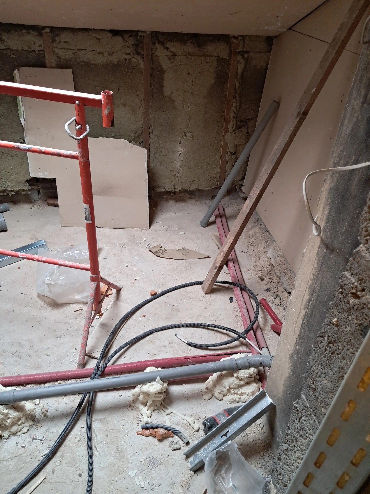 Remont w toku: widok na podłogę z rurami, przewodami, pianką montażową i rusztowaniem. Ściany w stanie surowym, płyty gipsowe częściowo zamontowane. Chaos remontowy.