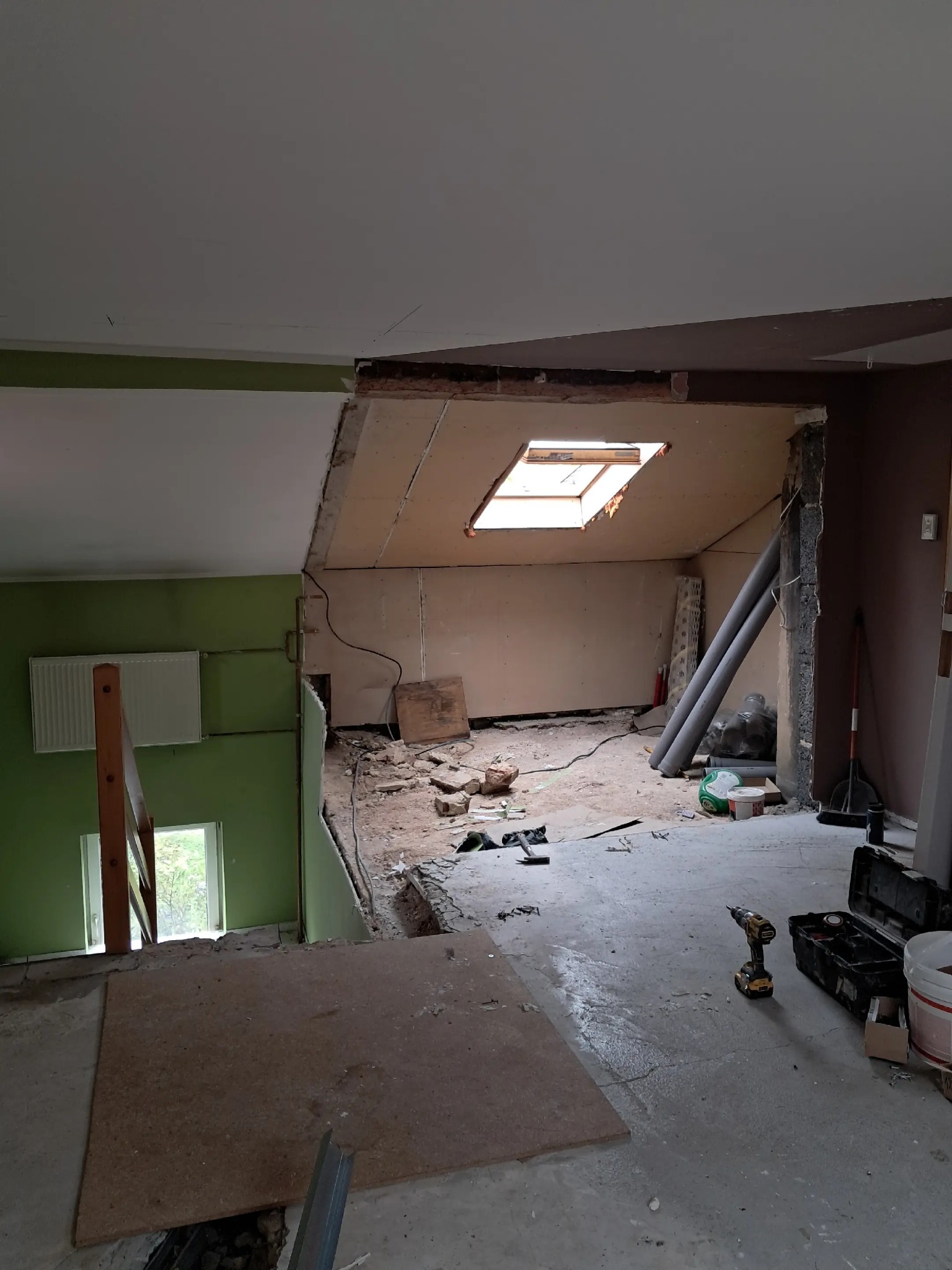 Wnętrze w trakcie przebudowy: widoczne odsłonięte ściany, okno dachowe, narzędzia i materiały budowlane. Fragment zielonej ściany z grzejnikiem.