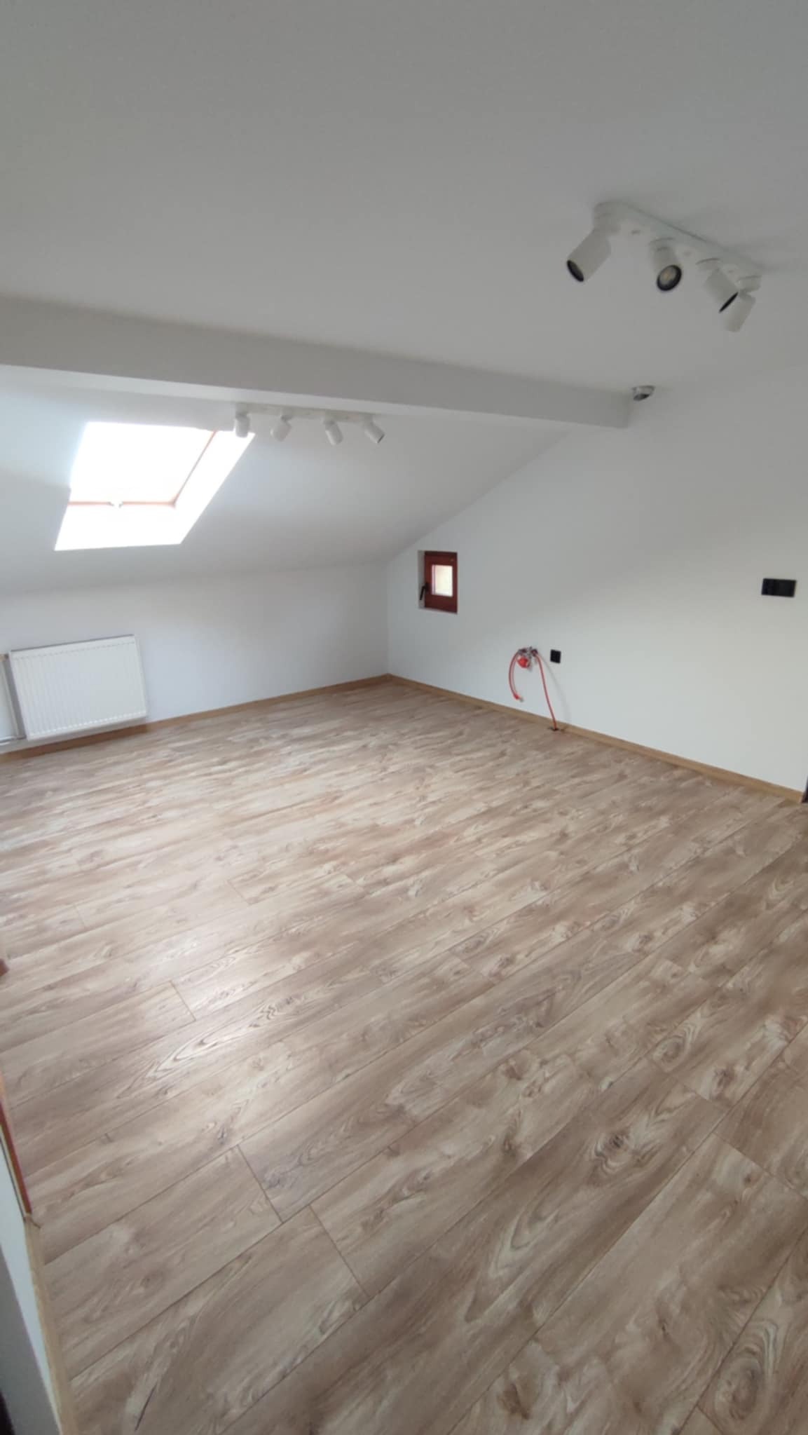 Jasne panele podłogowe w pokoju na poddaszu z oknem dachowym i białymi ścianami. Widoczne grzejnik i instalacja. Uwagę zwraca skośny sufit i oświetlenie.