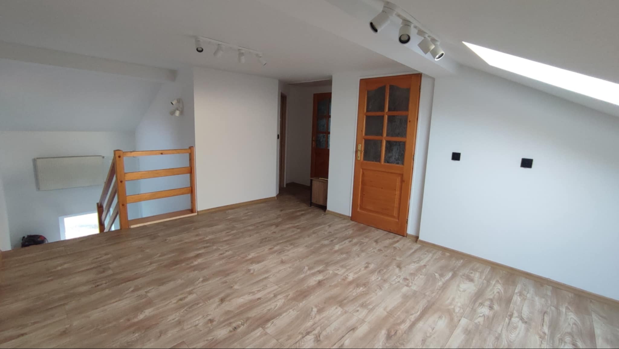 Jasne poddasze po remoncie: świeżo ułożone panele podłogowe w odcieniach drewna, białe ściany z oknem dachowym i drewniane drzwi z przeszkleniami.