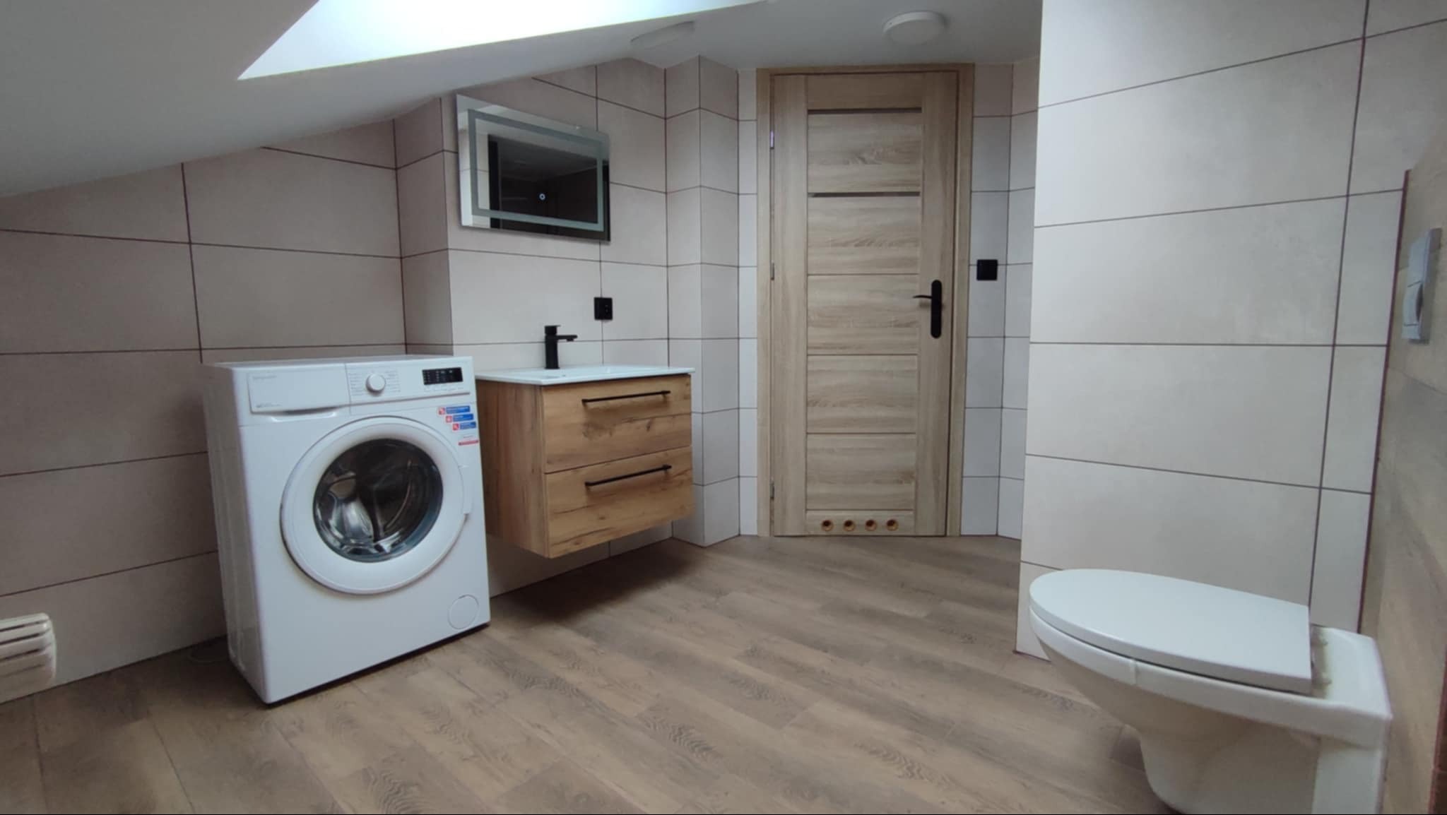 Jasna łazienka na poddaszu z pralką, drewnianą szafką pod umywalką i podświetlanym lustrem. Ukończone płytki na ścianach i drewnopodobna podłoga.