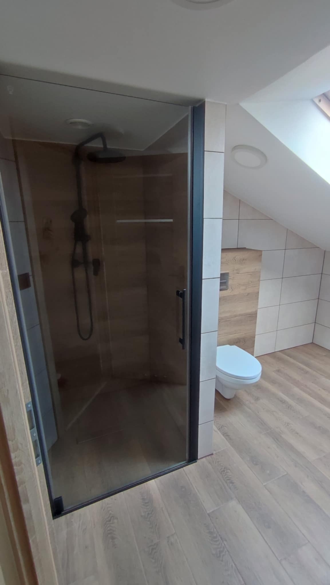 Wykończona łazienka na poddaszu ze szklaną kabiną prysznicową z czarnymi profilami, toaletą i drewnopodobnymi płytkami na podłodze i ścianach.