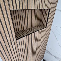 MJ BUD - Drewniana ściana z lamelami i wnęką z czarnym obramowaniem, obok marmurowe płytki podłogowe.