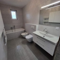 MJ BUD - Jasnoszara łazienka z mozaikową ścianą, białą armaturą, umywalką z szafką i podświetlanym lustrem, oświetlona kryształową lampą sufitową.