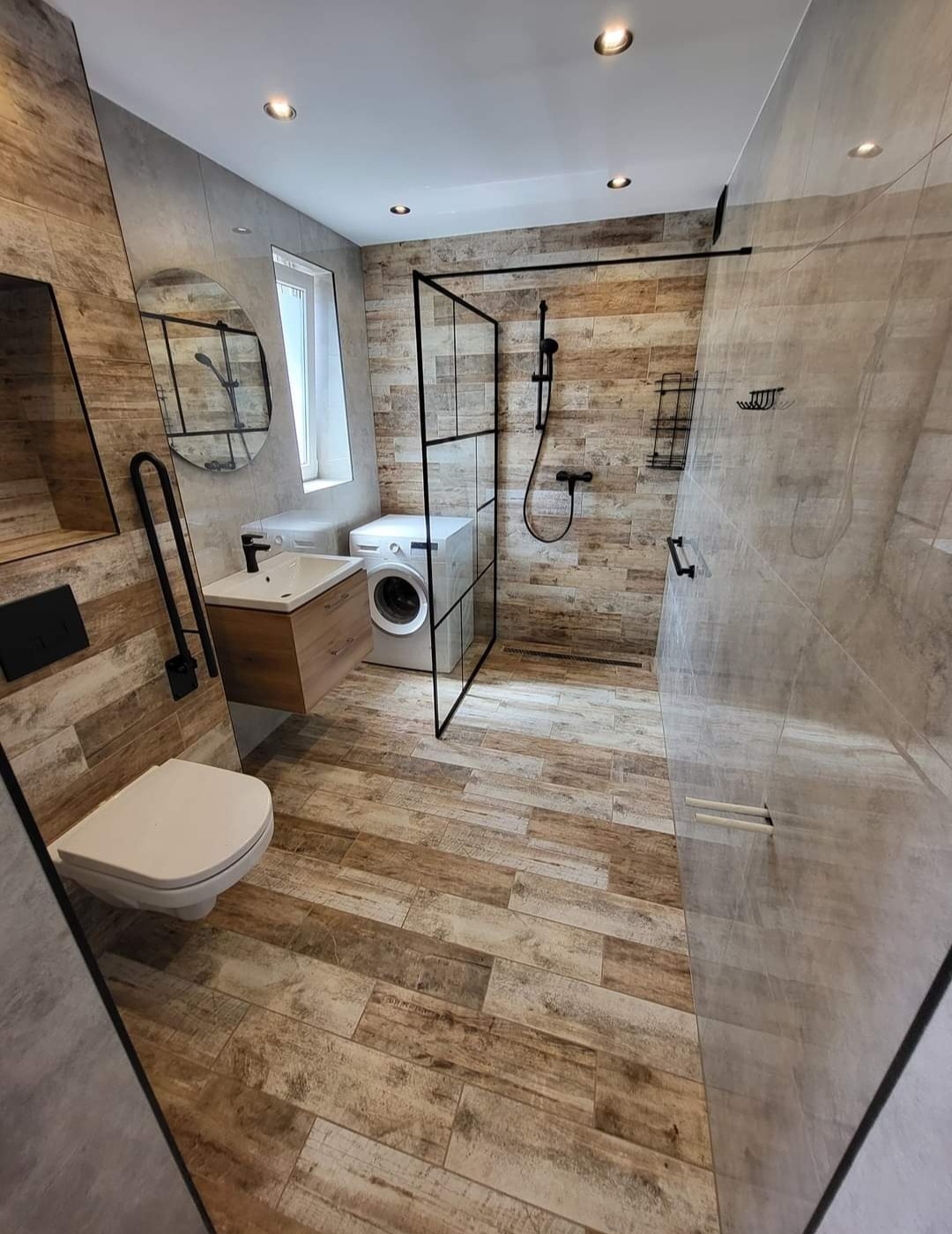 Nowoczesna łazienka z drewnopodobnymi płytkami, kabiną prysznicową typu walk-in z czarnymi ramami, wiszącą toaletą i pralką pod oknem.