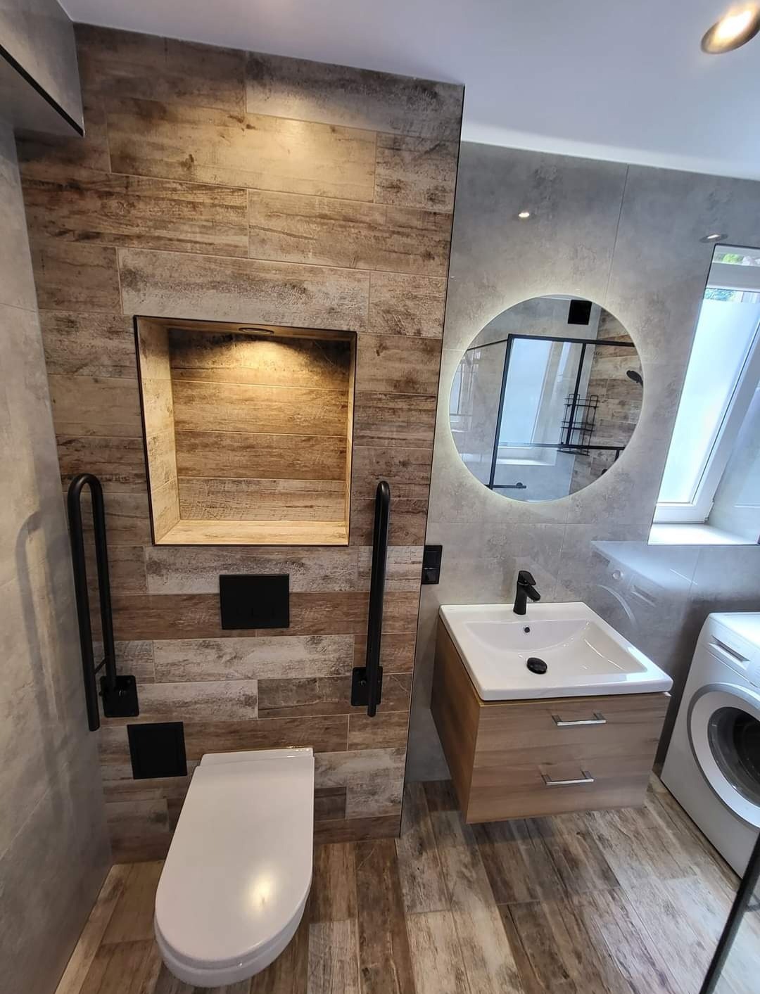 Nowoczesna łazienka z płytkami imitującymi drewno, okrągłym lustrem z podświetleniem LED, umywalką z szafką w kolorze drewna, toaletą z uchwytami dla niepełnosprawnych oraz wnęką w ścianie...