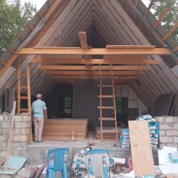 Sakhavat Samadov - Wnętrze poddasza w trakcie adaptacji: widoczne drewniane krokwie, izolacja, stosy płyt OSB, worki z zaprawą i drabina; osoba wchodzi do pomieszczenia.