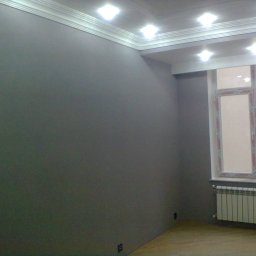 Sakhavat Samadov - Ściana pomalowana na szaro z białym gzymsem sufitowym i grzejnikiem pod oknem, widok na budynek z zewnątrz.