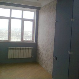 Sakhavat Samadov - Pokój po odświeżeniu: ściana z wzorzystą tapetą, okno z widokiem na miasto, grzejnik i ciemnoszara szafa z zabezpieczeniami na drzwiach.