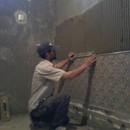 Sakhavat Samadov - Mężczyzna w czapce, kuca, poziomuje wzorzyste płytki na ścianie pokrytej klejem, widoczna kielnia i gniazdko elektryczne.