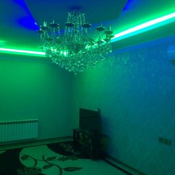 Sakhavat Samadov - Kryształowy żyrandol podświetlony zielonymi i niebieskimi neonami w pokoju z wzorzystą tapetą, widoczny fragment grzejnika i dywanu z kwiatowym motywem.