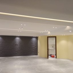 Sakhavat Samadov - Wnętrze po remoncie: szara ceglana ściana, jasna podłoga z płytek, żółta ściana z oświetleniem LED, sufit z punktowym oświetleniem. Widoczny fragment kuchni z białymi szafkami.