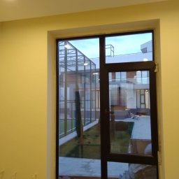 Sakhavat Samadov - Ciemnobrązowe okno z widokiem na nowoczesny budynek z szklaną konstrukcją i zielonym trawnikiem, widoczne materiały budowlane w dolnej części okna wewnątrz pomieszczenia.