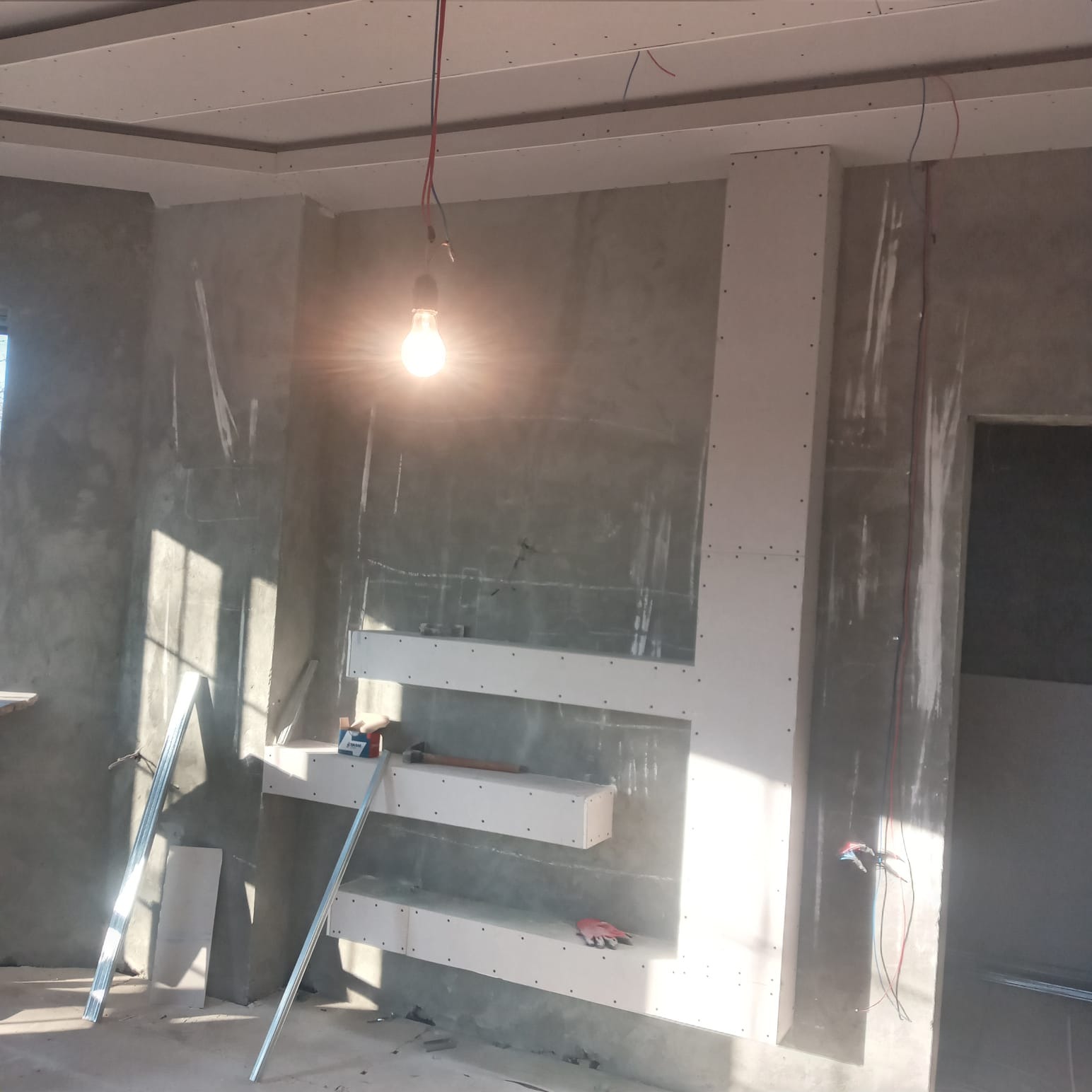 Wnętrze w trakcie remontu z konstrukcją z płyt kartonowo-gipsowych na ścianie, widoczne narzędzia i wisząca żarówka.