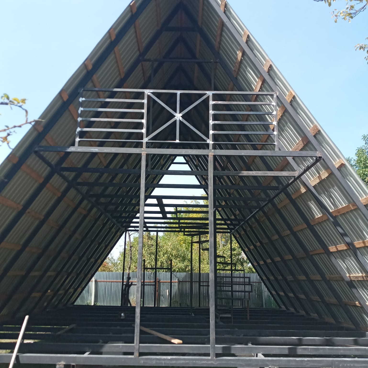 Szkielet konstrukcji domu w kształcie litery A z metalowymi belkami, widok z wnętrza na zewnątrz, z widoczną konstrukcją dachu z blachy falistej i drewnianych łat, oraz metalową konstrukcją antresoli.