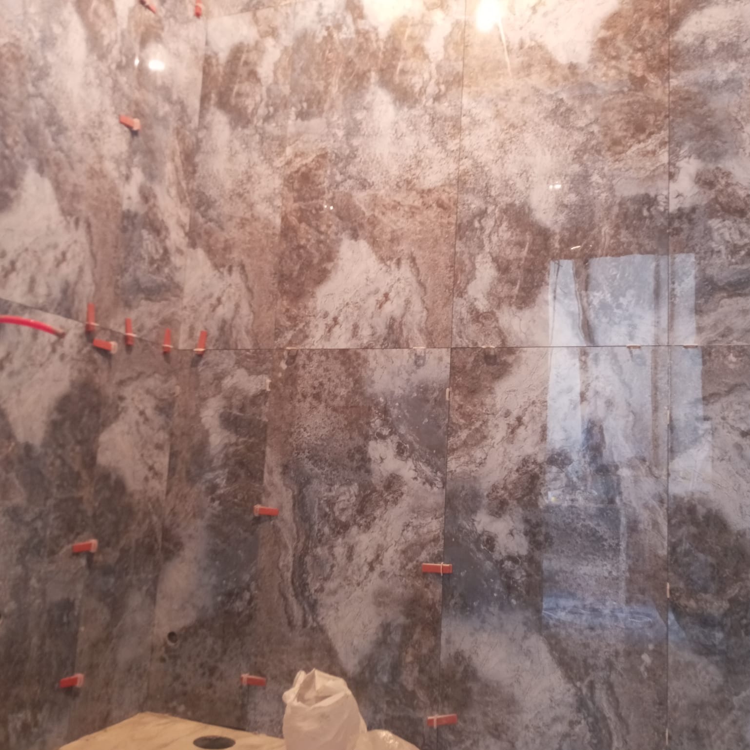 Ściana łazienki w trakcie układania płytek imitujących marmur, z widocznymi krzyżykami dystansowymi i workiem z zaprawą klejową w dolnym rogu.