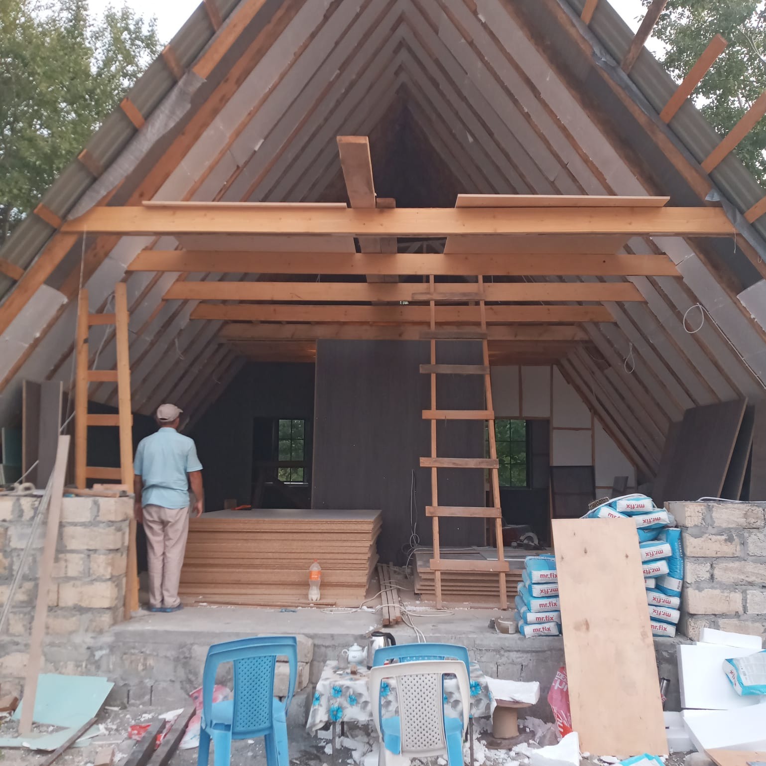 Wnętrze poddasza w trakcie adaptacji: widoczne drewniane krokwie, izolacja, stosy płyt OSB, worki z zaprawą i drabina; osoba wchodzi do pomieszczenia.