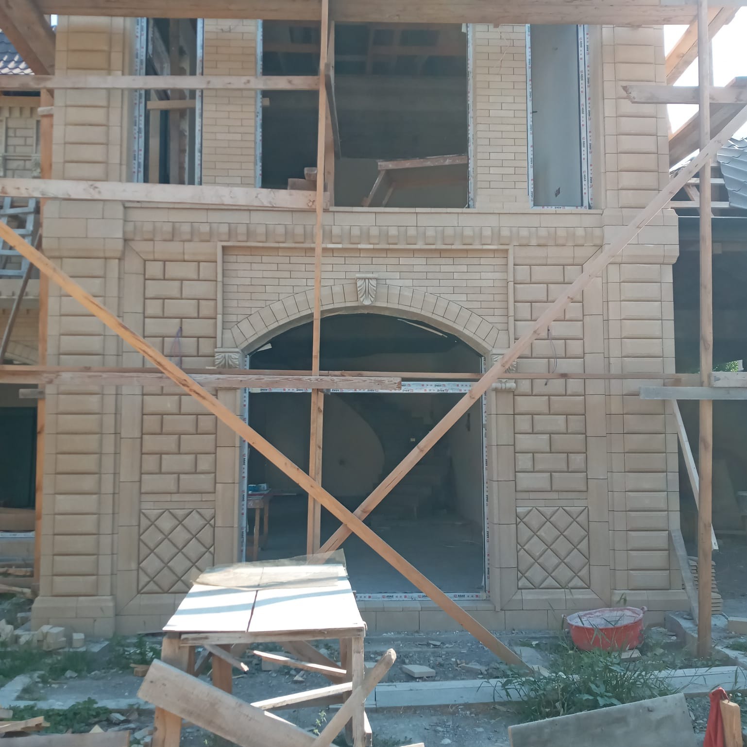 Budynek w trakcie wznoszenia elewacji z dekoracyjnej, beżowej cegły, widoczne rusztowania i niezabudowane otwory okienne.