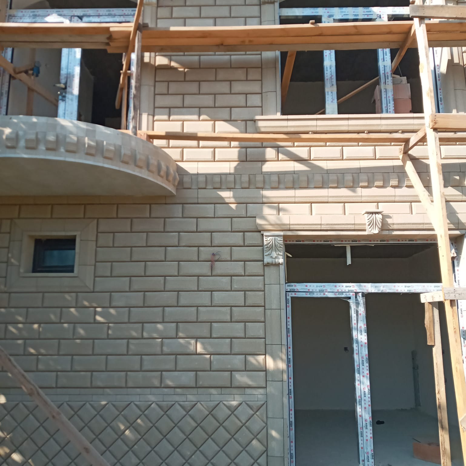 Elewacja budynku w trakcie wykończenia z beżowej cegły klinkierowej, widoczne rusztowania i przygotowane otwory okienne oraz drzwiowe z zamontowaną stolarką okienną, dekoracyjne elementy...