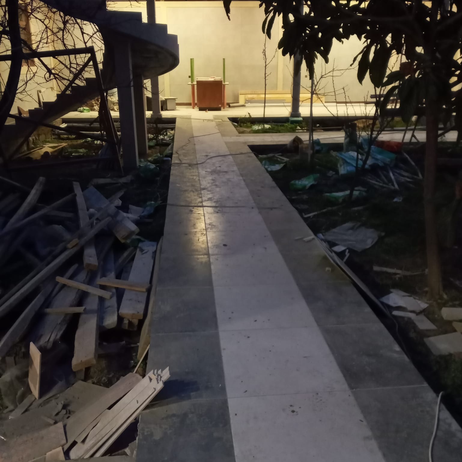 Układanie płytek chodnikowych na zewnątrz budynku z elementami spiralnych schodów i pozostałościami po pracach budowlanych, widoczne deski i gruz.