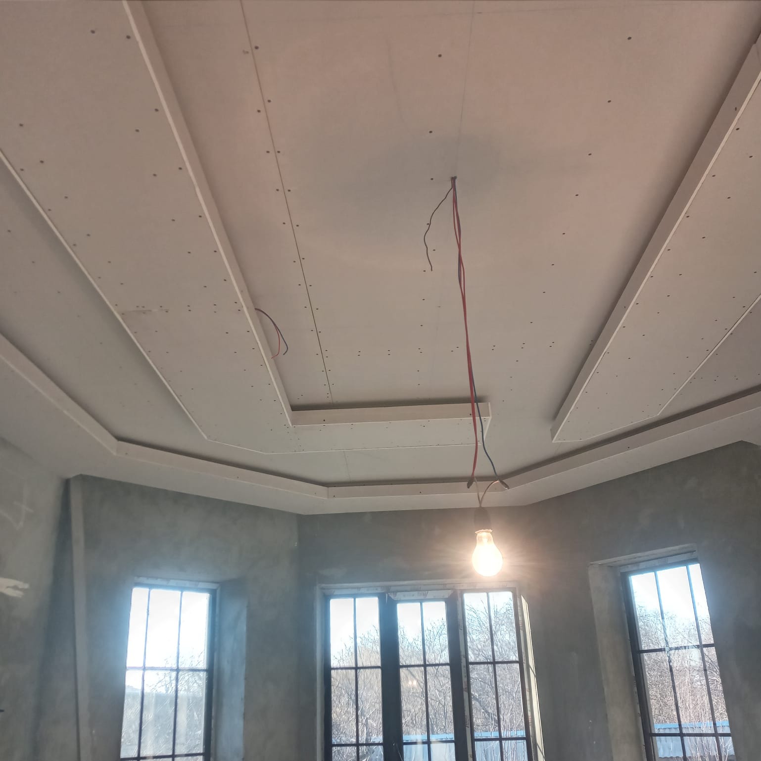 Sufit podwieszany z płyt kartonowo-gipsowych w trakcie montażu, widoczne profile i kable elektryczne, surowe ściany w tle, oświetlone żarówką, trzy okna z widokiem na drzewa bez liści.