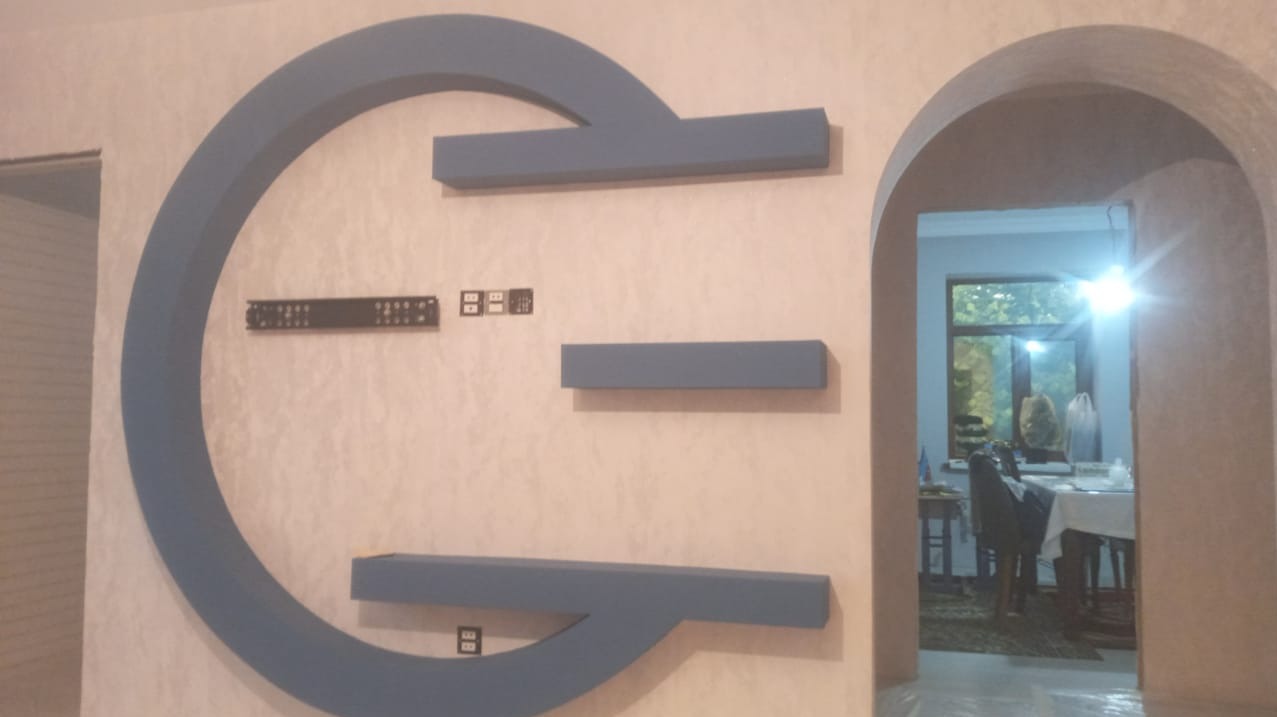 Ściana z dekoracyjną, niebieską konstrukcją przypominającą literę G, z trzema półkami i gniazdkami elektrycznymi, widok na pokój przez łukowate przejście.