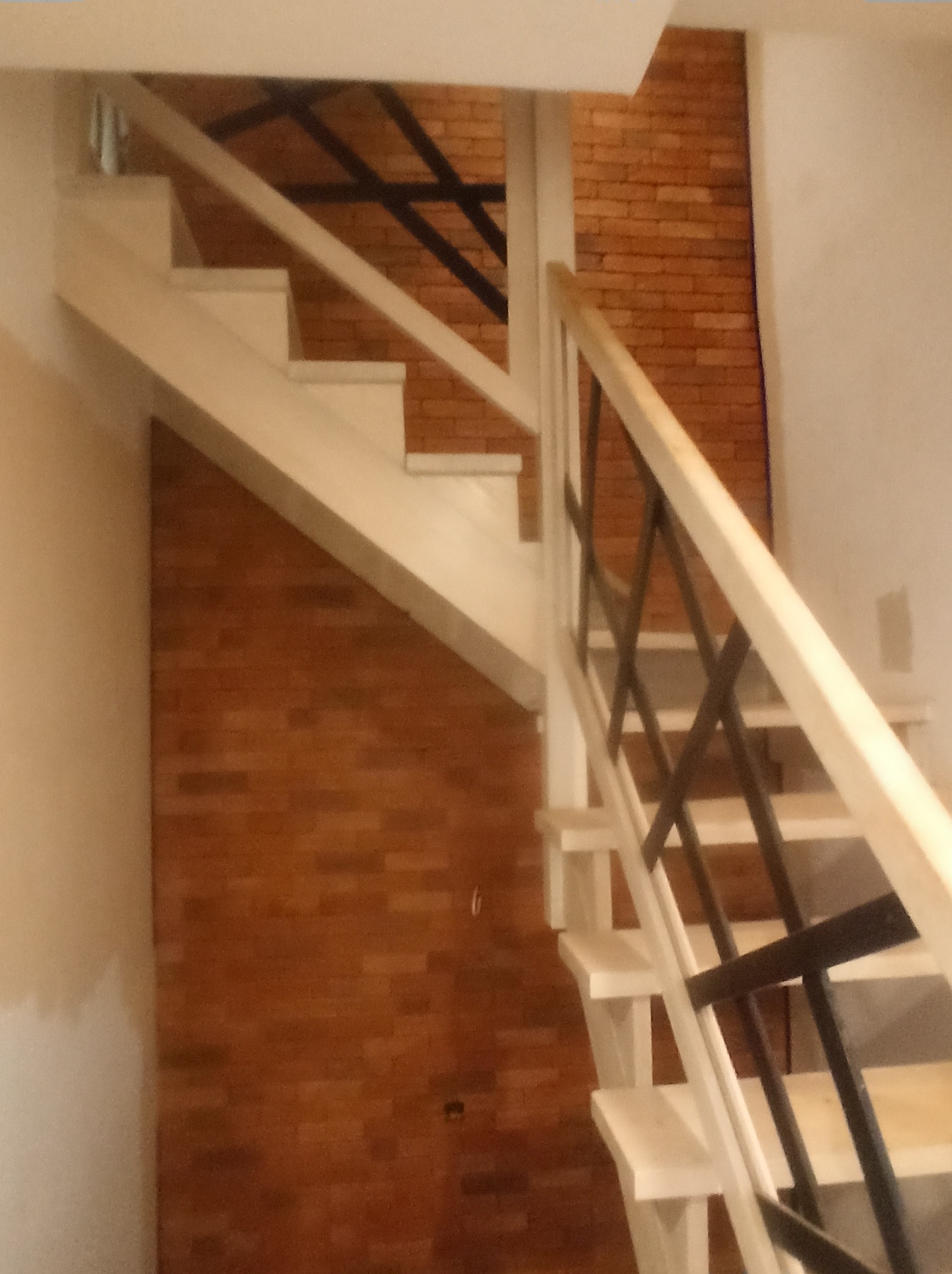 Nowe, jasne schody z drewnianą balustradą i czarnymi, metalowymi akcentami na tle ściany wyłożonej cegłą.