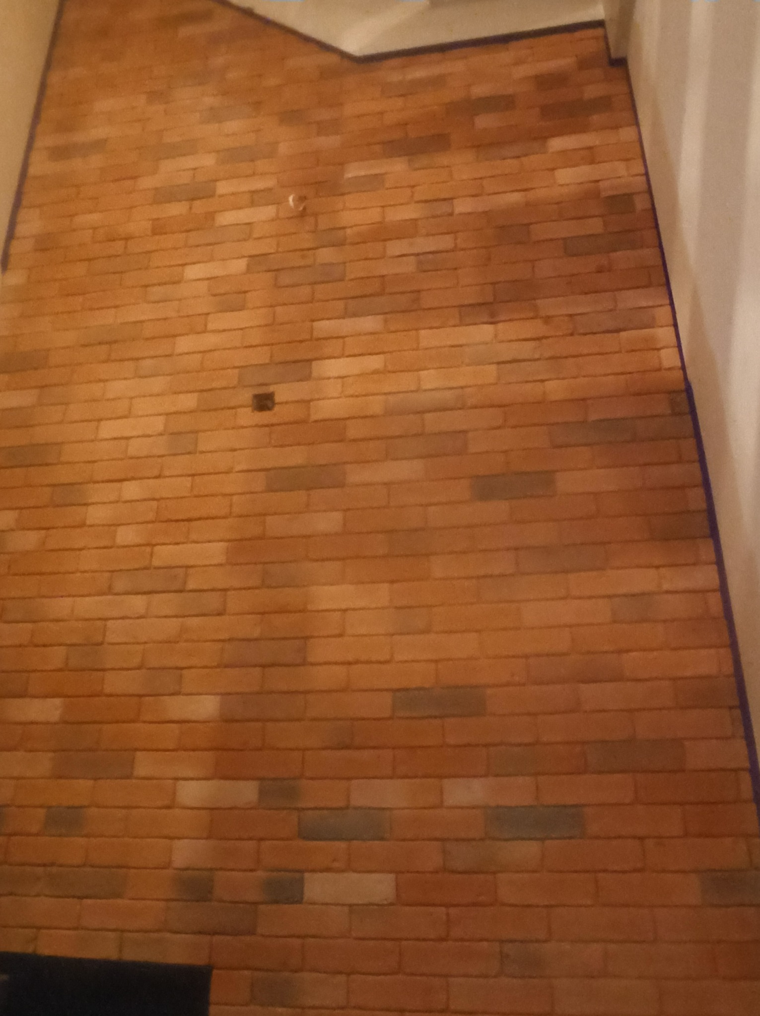 Ściana z cegły klinkierowej w odcieniach brązu i szarości, z widocznym otworem na gniazdko elektryczne i delikatnym oświetleniem padającym z góry.