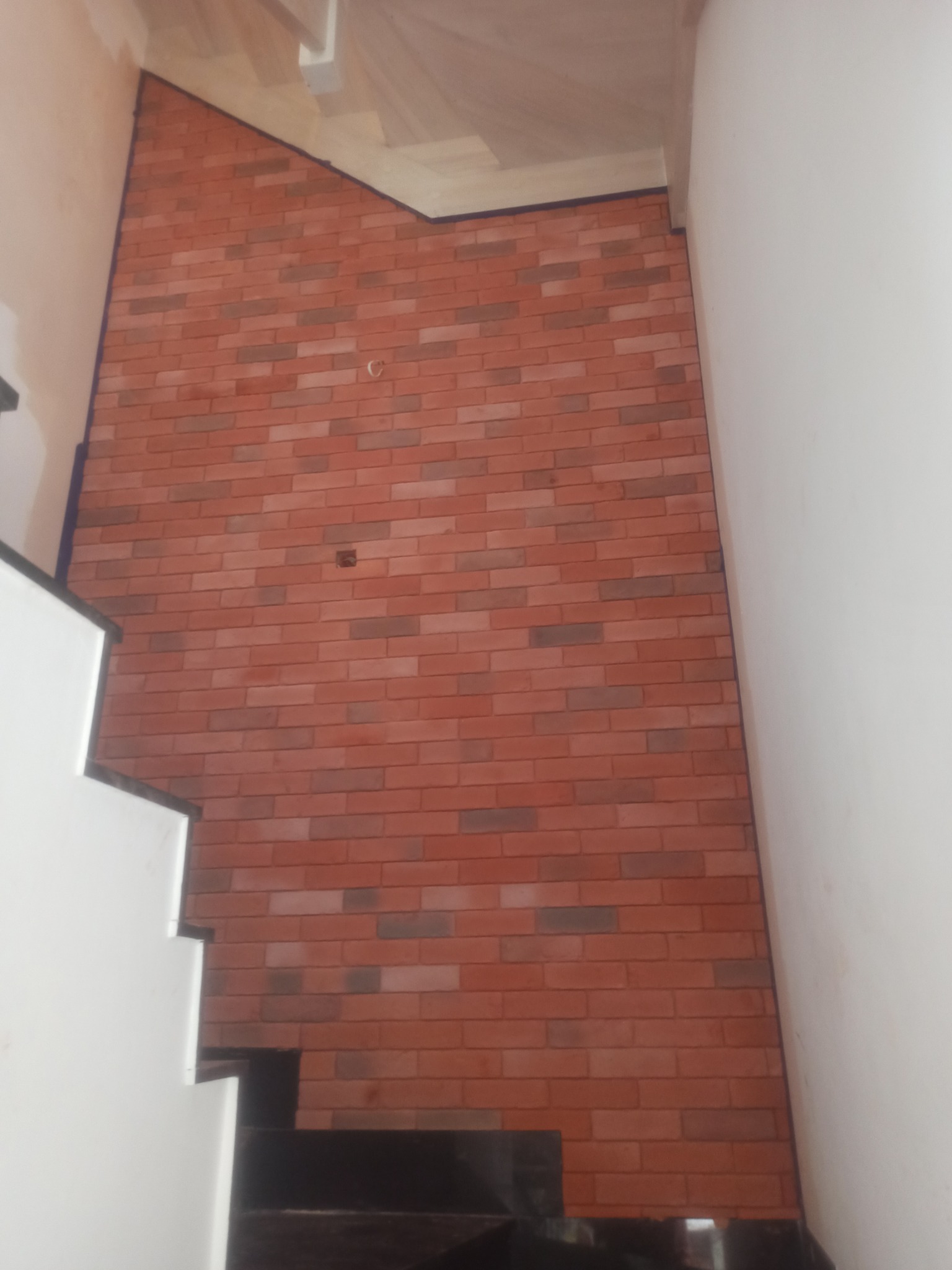 Ściana wykończona czerwoną cegłą klinkierową w korytarzu przy schodach, widoczny fragment schodów z białymi stopniami i ciemnym podstopniem.