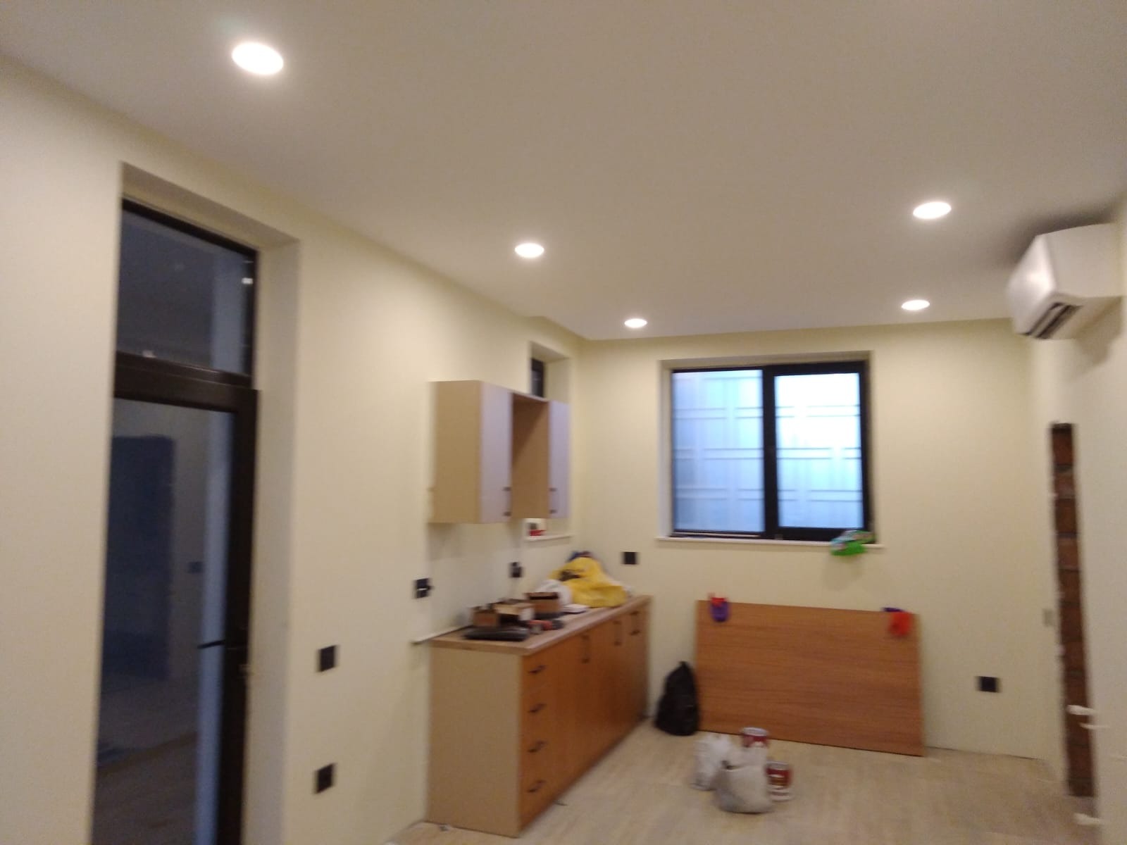 Wnętrze pokoju z przygotowaną ścianą pod malowanie, widoczne puszki farb i narzędzia malarskie, zamaskowane okno, surowy drewniany blat oraz szafki kuchenne bez frontów.