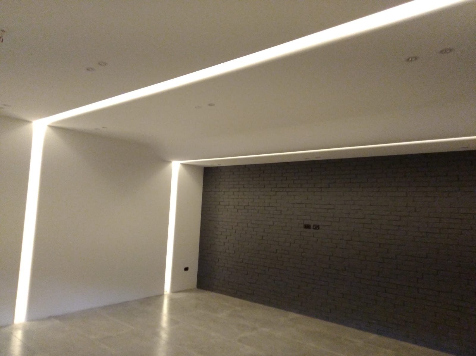 Pokój z nowoczesnym oświetleniem LED w listwach wzdłuż sufitu i ścian, szara ściana z cegły, jasne ściany i podłoga.