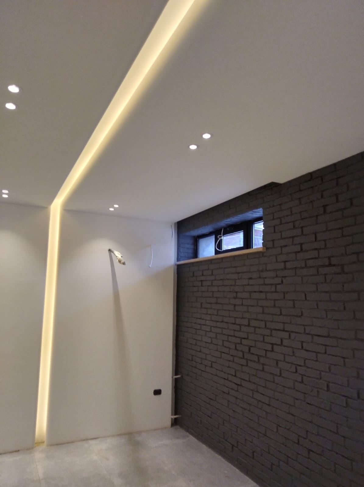 Nowoczesne wnętrze z szarą ceglaną ścianą, oświetleniem LED w suficie i punktowymi lampami, małym oknem z drewnianym parapetem i szarą podłogą.