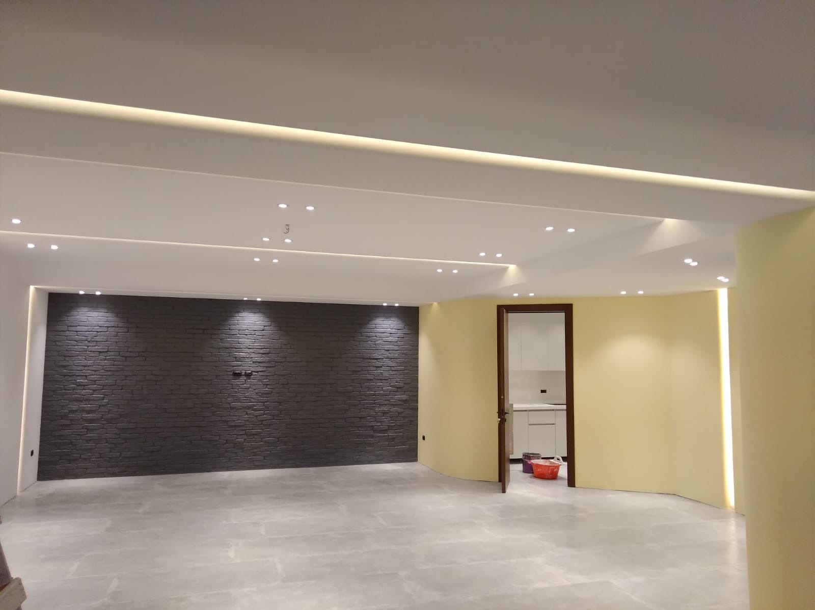 Wnętrze po remoncie: szara ceglana ściana, jasna podłoga z płytek, żółta ściana z oświetleniem LED, sufit z punktowym oświetleniem. Widoczny fragment kuchni z białymi szafkami.
