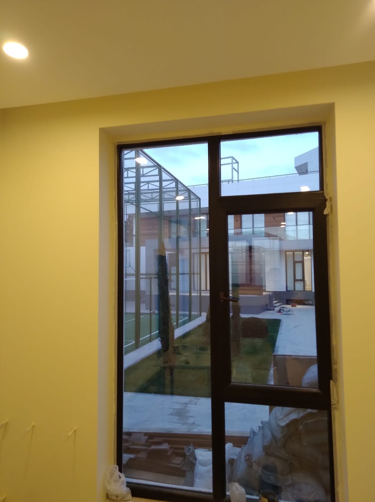 Ciemnobrązowe okno z widokiem na nowoczesny budynek z szklaną konstrukcją i zielonym trawnikiem, widoczne materiały budowlane w dolnej części okna wewnątrz pomieszczenia.