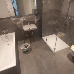 Alpeks.sp.zoo - Nowoczesna łazienka w trakcie wykańczania: widoczna wanna, umywalka zakryta folią, kabina prysznicowa z szarymi płytkami i wiadro z narzędziami Brilux na podłodze.