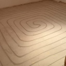 Alpeks.sp.zoo - Rury spiralnie ułożone w wylewce podłogowej, przygotowane do montażu systemu grzewczego w jasnym pomieszczeniu z surowymi ścianami.