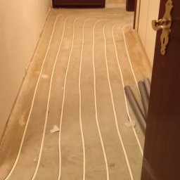 Alpeks.sp.zoo - Białe rurki systemu ogrzewania podłogowego ułożone na szarej wylewce w korytarzu, widoczne rury przy ścianie i fragment otwartych drzwi z mosiężną klamką.