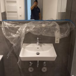 Alpeks.sp.zoo - Nowa, biała umywalka zamontowana na szarych płytkach, zabezpieczona folią malarską na czas remontu łazienki, widoczne przyłącza i bateria, odbicie osoby robiącej zdjęcie w lustrze.