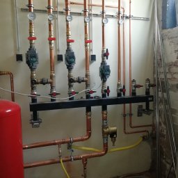 Alpeks.sp.zoo - Rozdzielacz ogrzewania podłogowego z miedzianymi rurami, manometrami i zaworami termostatycznymi na tle surowej ściany w trakcie montażu.