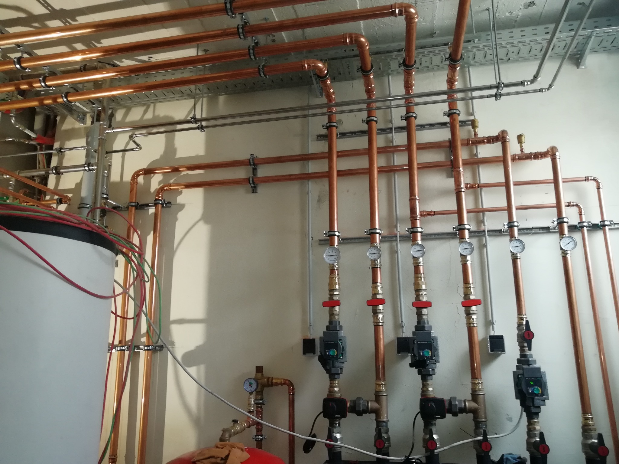 Rozbudowana instalacja z rur miedzianych z zaworami, manometrami i pompami obiegowymi, zamontowana na ścianie w pomieszczeniu technicznym.