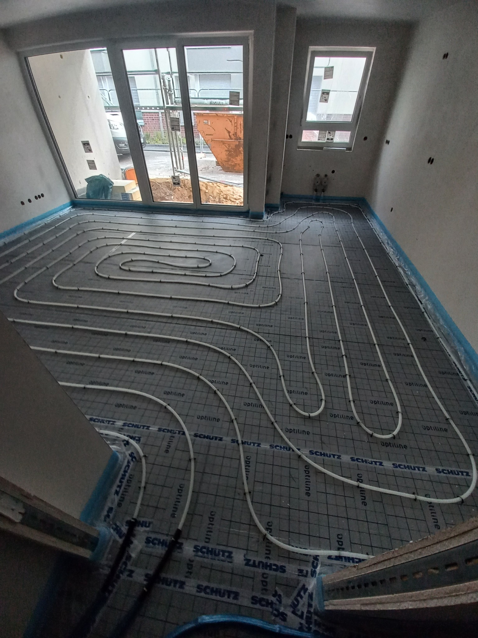 Rozłożone rury systemu grzewczego na podłodze z matą systemową w surowym wnętrzu, widok z góry na instalację przed wylewką, rury marki Optiline i mata Schutz.