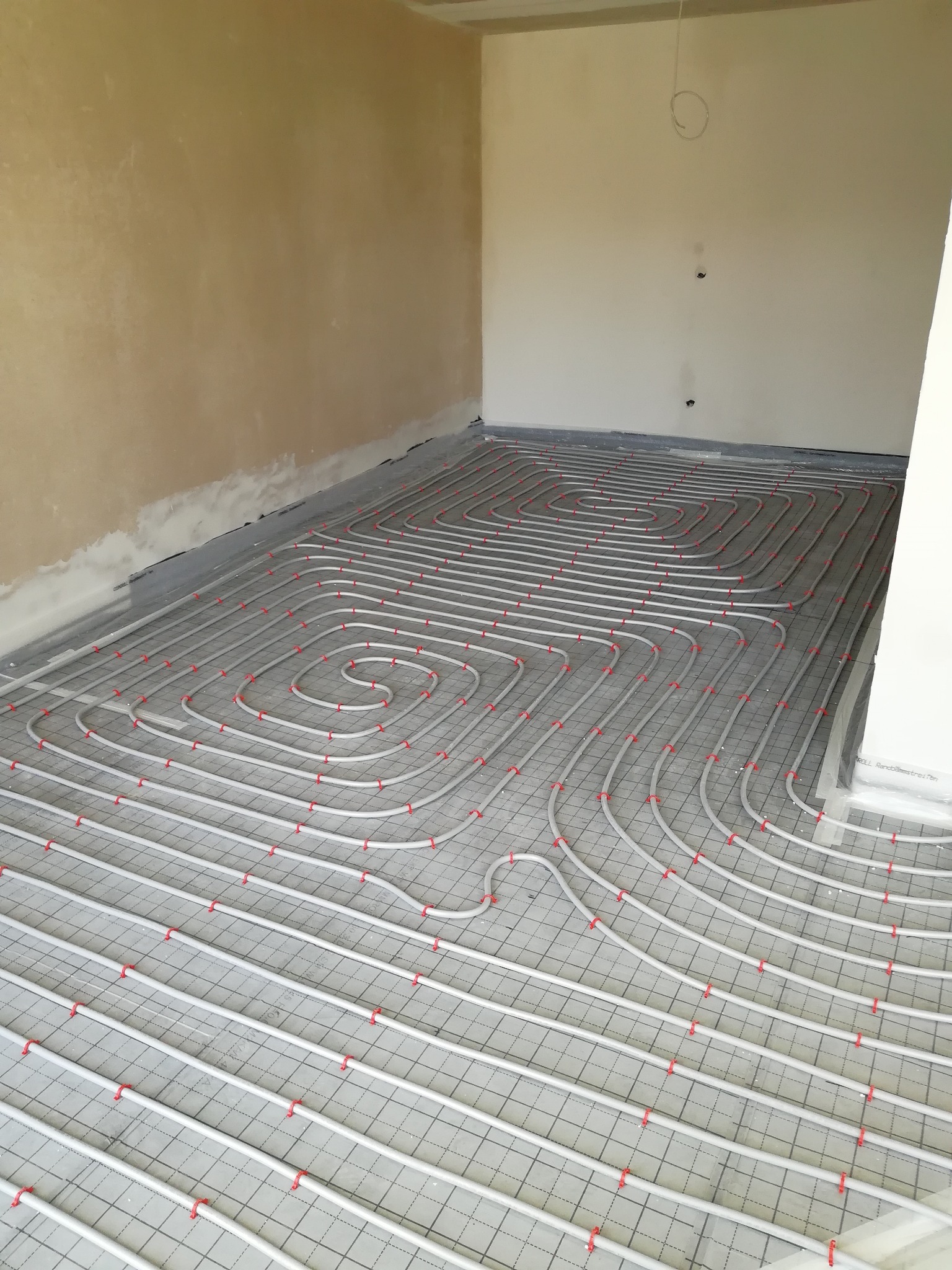 Rozłożone rury grzewcze na podkładzie z siatki w surowym pomieszczeniu, przygotowane do zalania wylewką.