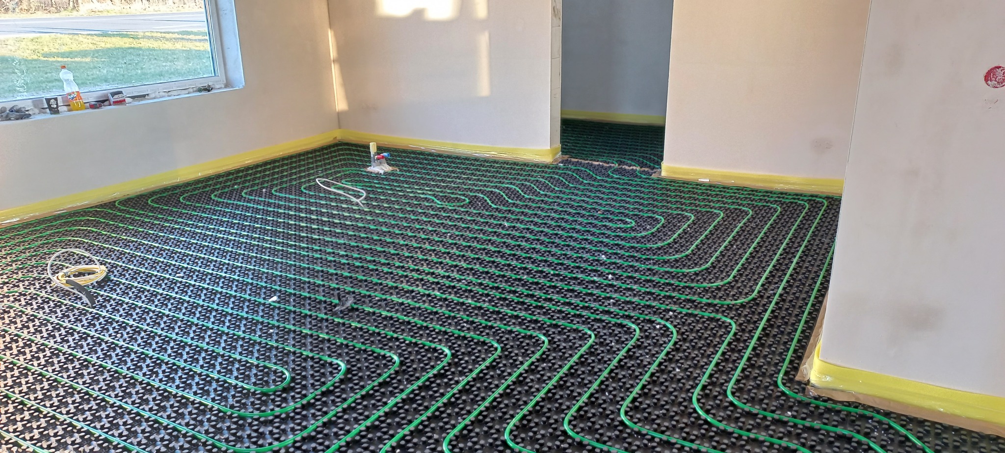 Rozłożona instalacja ogrzewania podłogowego z zielonych rurek na czarnej macie, zabezpieczona żółtą taśmą malarską przy ścianach, widok na sąsiednie pomieszczenie z identycznym systemem.