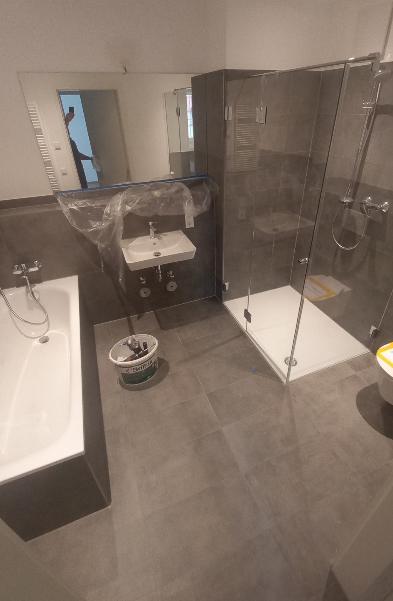 Nowoczesna łazienka w trakcie wykańczania: widoczna wanna, umywalka zakryta folią, kabina prysznicowa z szarymi płytkami i wiadro z narzędziami Brilux na podłodze.