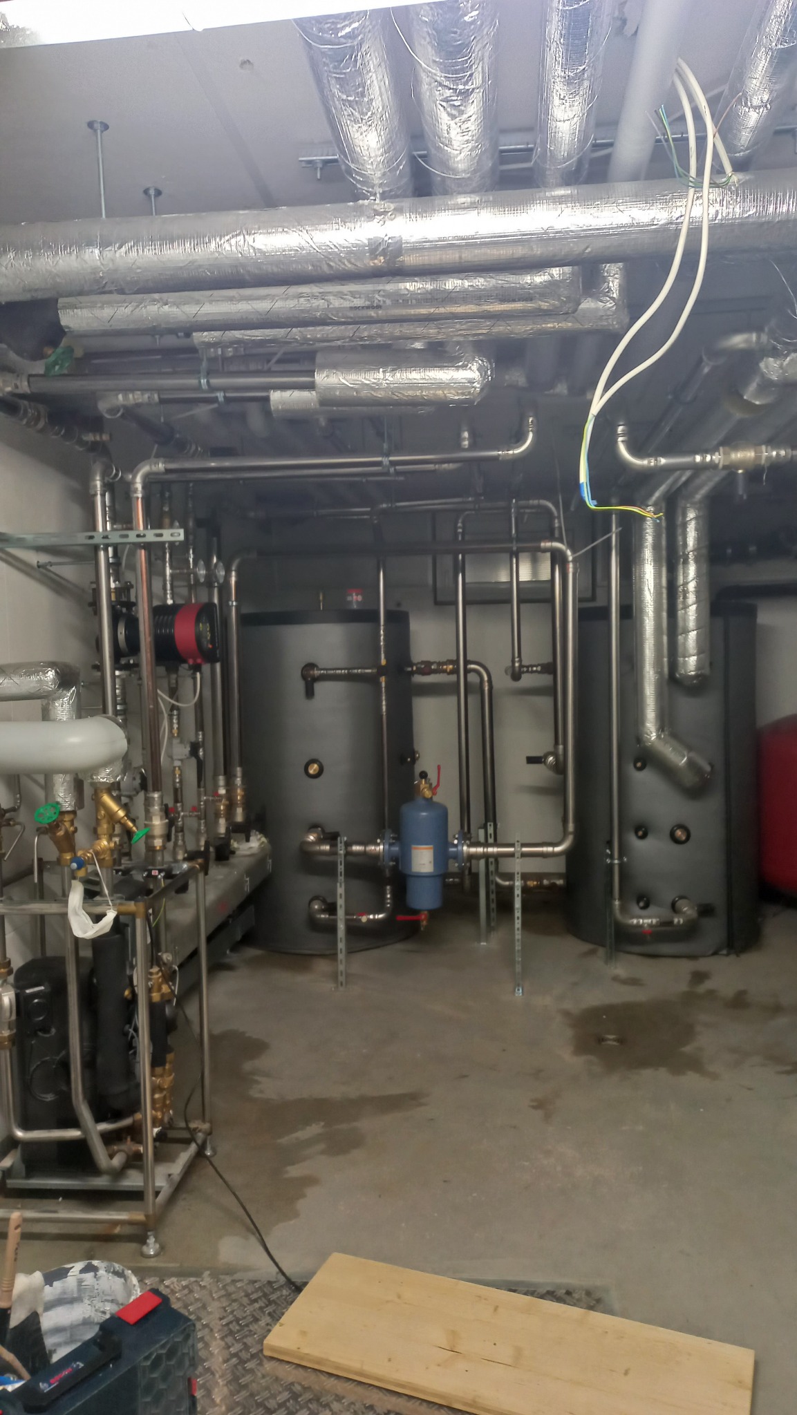 Rozbudowana instalacja grzewcza w piwnicy, widoczne zbiorniki akumulacyjne, rury, zawory, izolacja termiczna na rurach pod sufitem, narzędzia na podłodze.