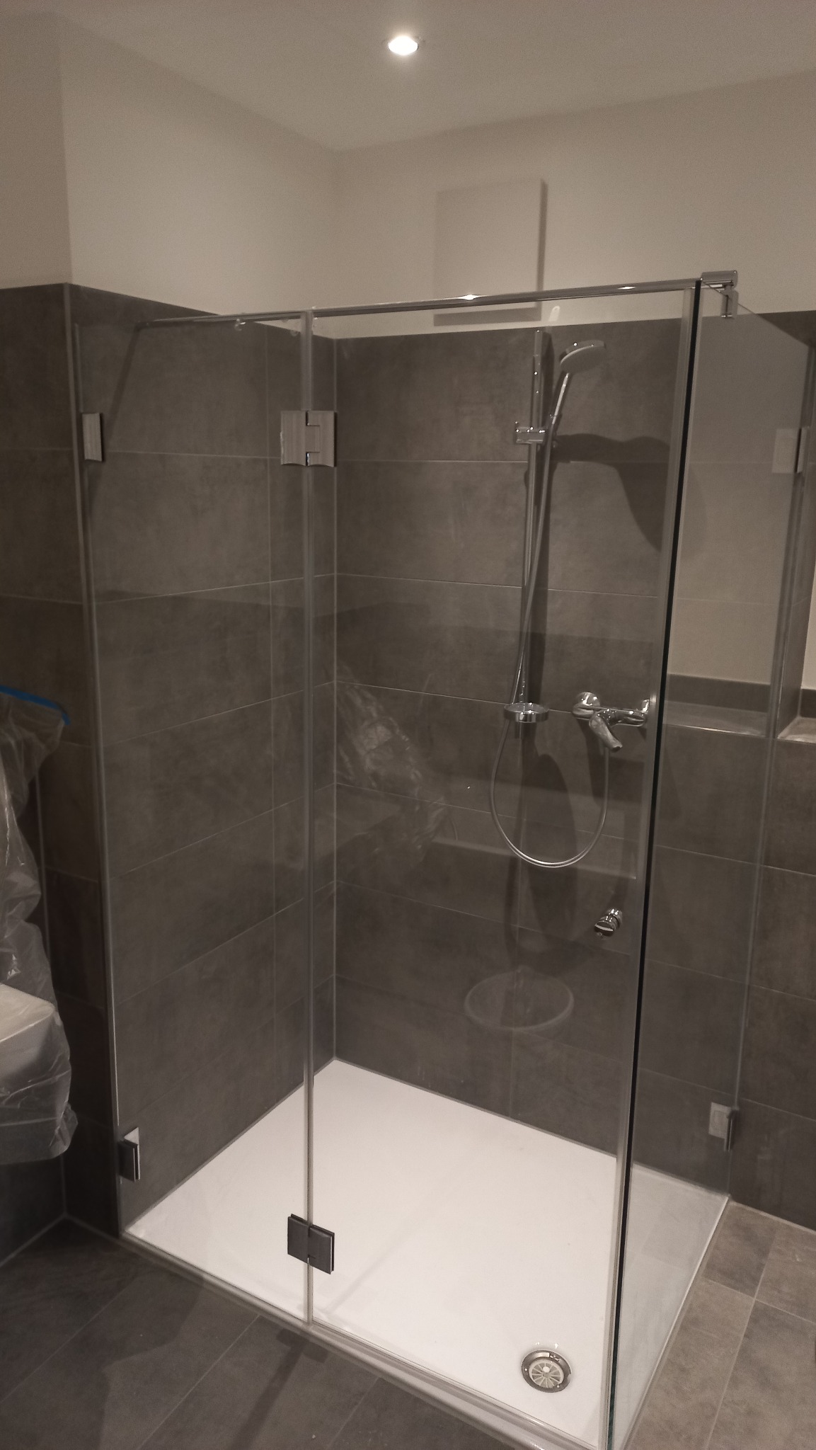 Nowoczesna kabina prysznicowa z ciemnoszarymi płytkami, chromowanymi okuciami i białą brodzikiem, widok z zewnątrz, częściowo zakryta folią ochronną po instalacji.